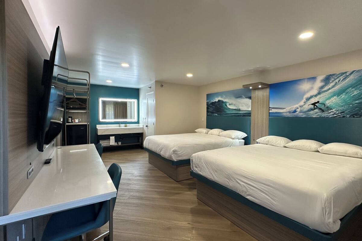 A hotel room with two beds, a desk, a TV, and a large ocean wave mural on the wall.