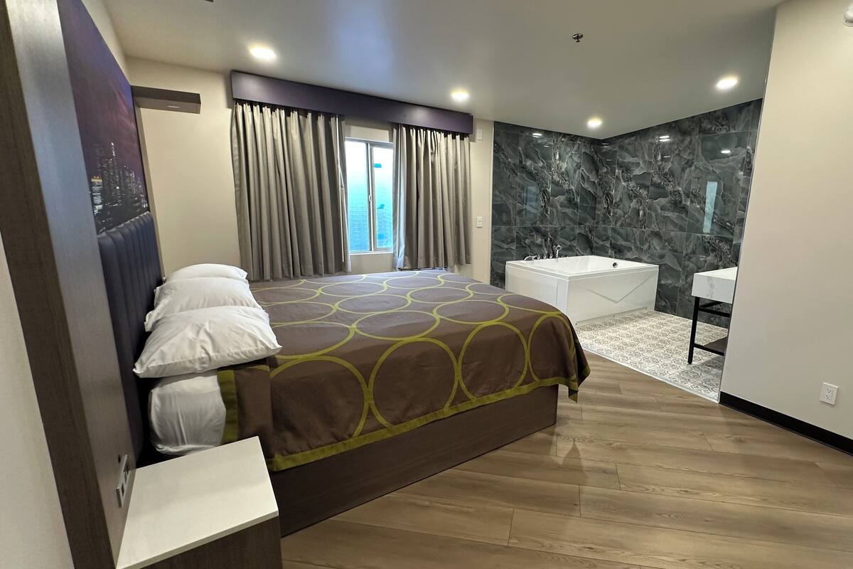 A modern hotel room with a large bed, a separate bathroom with a bathtub, and a view of the city through the window.
