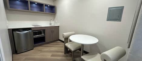 A compact kitchen with a built-in microwave, a stainless steel refrigerator, and a sink. There is a round table with two chairs and a small cabinet above the counter.