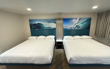 A hotel room with two beds, a large ocean wave painting on the wall, and a window with curtains.