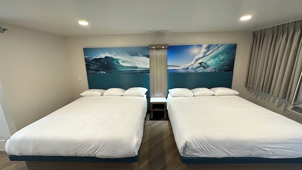 A hotel room with two beds, a large ocean wave painting on the wall, and a window with curtains.