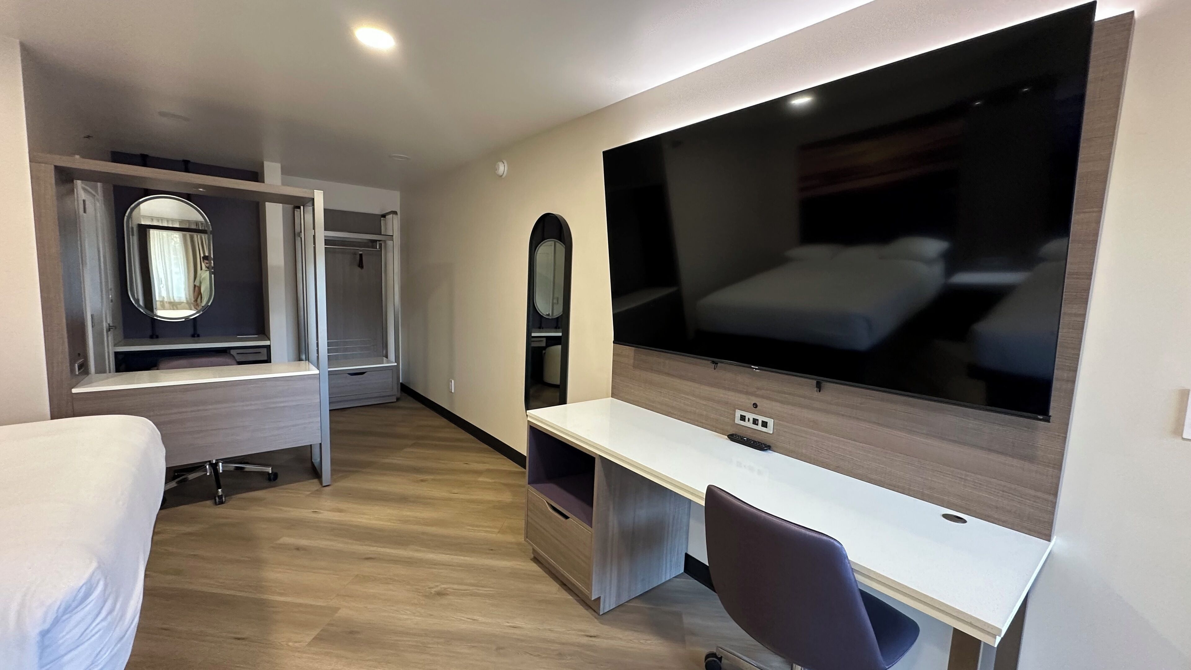 A modern hotel room with a large bed, a desk with a chair, a TV, a wardrobe with a mirror, and a small seating area.