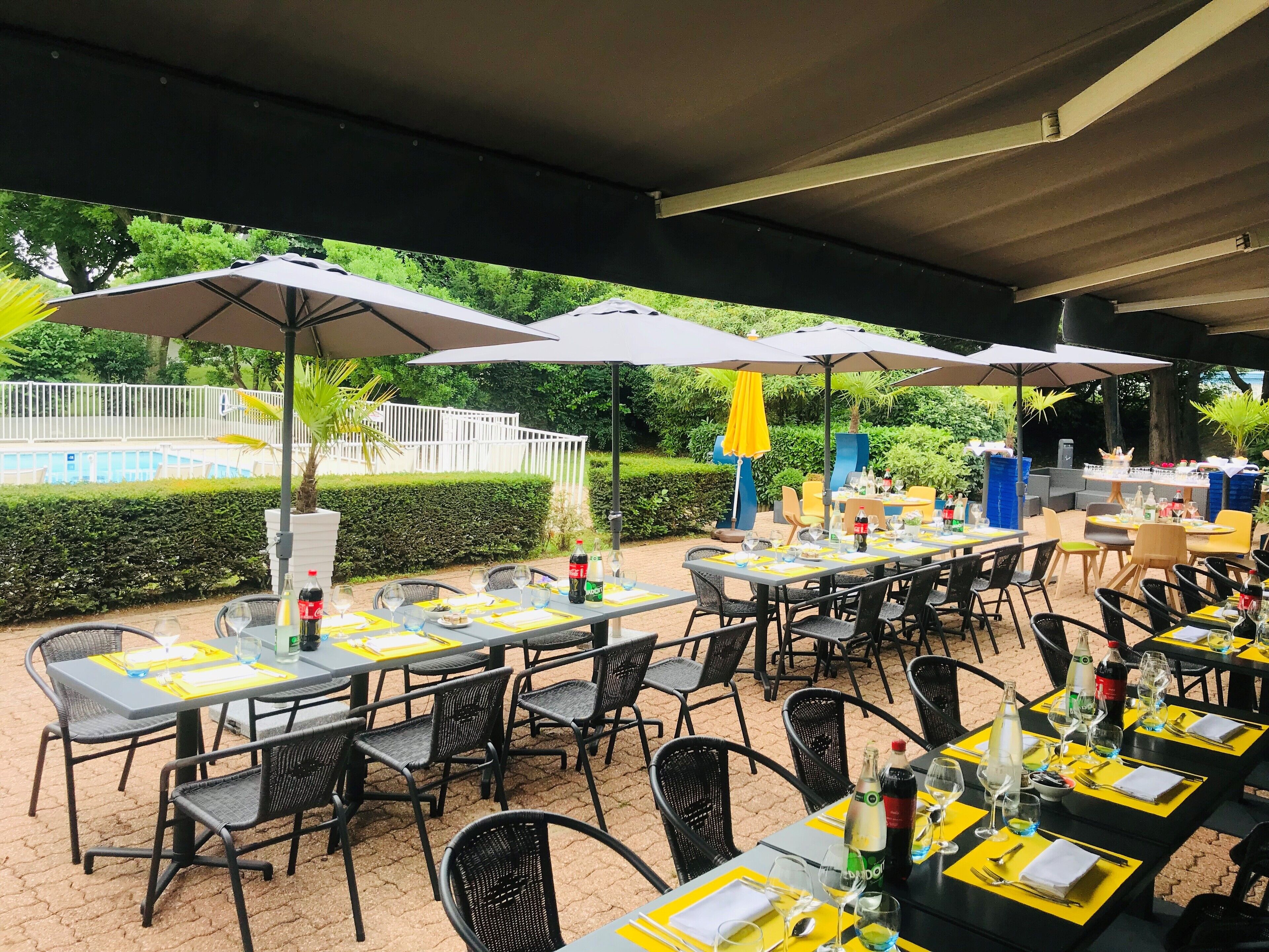 An outdoor dining area with tables and chairs, umbrellas, and a pool in the background.