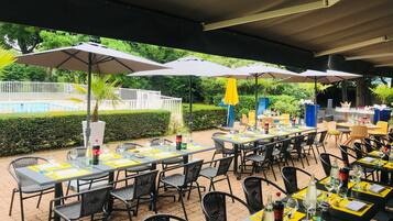 Un área de comedor al aire libre con mesas y sillas, sombrillas y una piscina al fondo.