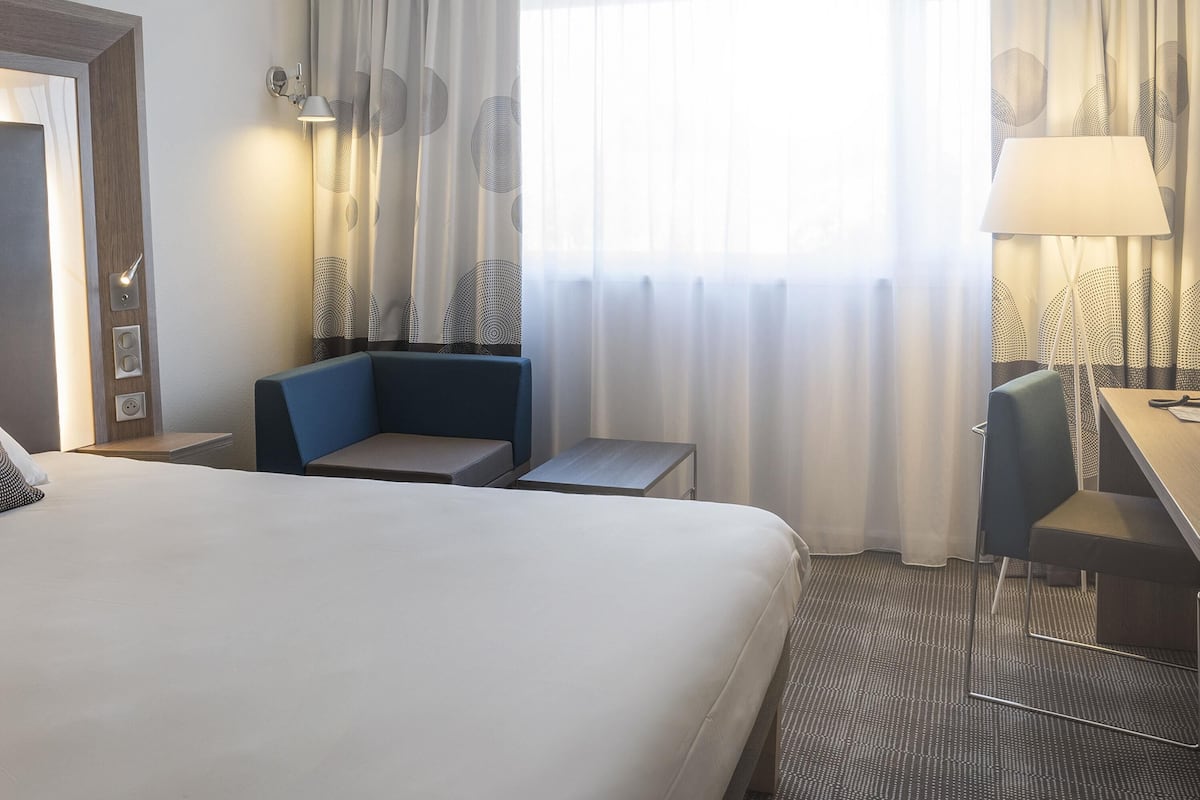 A hotel room with a large bed, a desk, a chair, a small table, and a sofa. There are curtains on the window and a lamp on the desk.