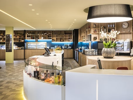 Ein moderner Barbereich mit einer Glastheke, Regalen mit Flaschen, einer Uhr und einer großen Vase mit Blumen.. Novotel Paris Nord Expo Aulnay Hotel