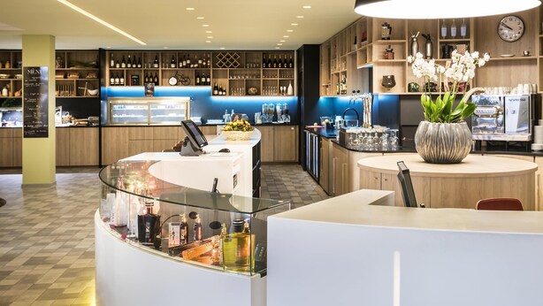 A modern bar area with a glass counter, a Nespresso machine, and a variety of bottles and glasses. - Novotel Paris Nord Expo Aulnay Hotel (Aulnay-sous-Bois)