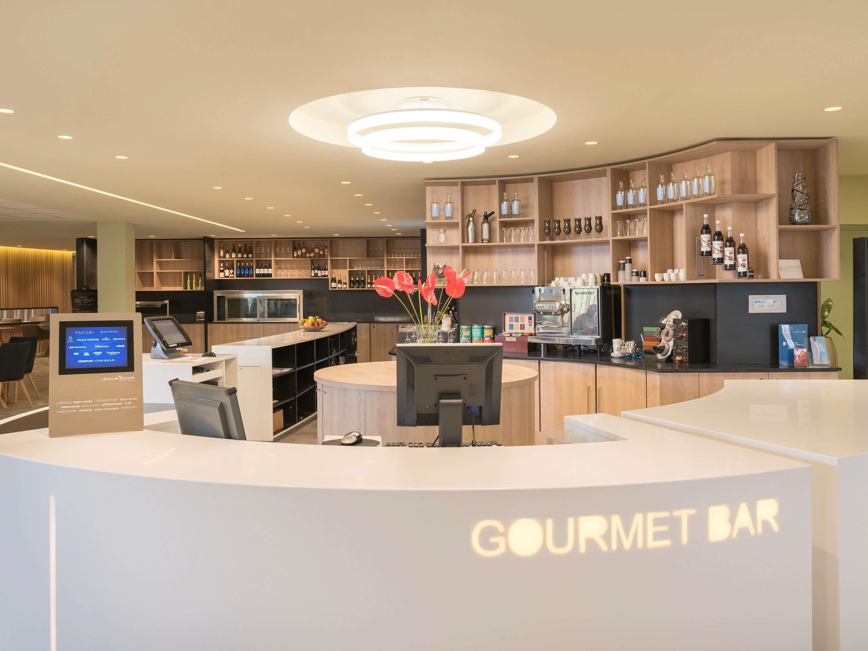 Un espace bar moderne doté d’un écran numérique affichant le menu, d’une machine à café, ainsi que de diverses bouteilles et verres présentés sur des étagères.