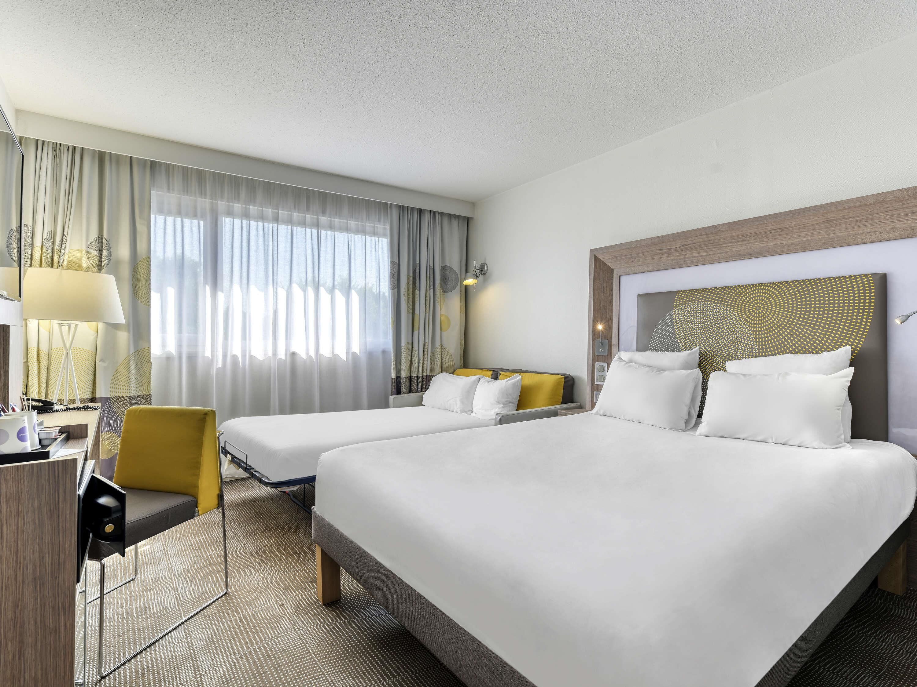 A hotel room with two beds, a desk, a chair, and a large window with curtains.