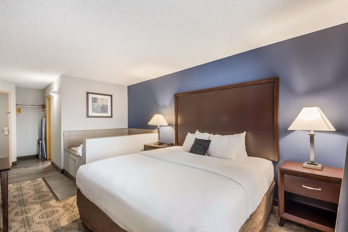 Une chambre d’hôtel avec un grand lit, deux tables de chevet avec des lampes, une tête de lit en bois, un tableau encadré au mur et une salle de bain équipée d’un porte-serviettes et d’un rideau de douche.