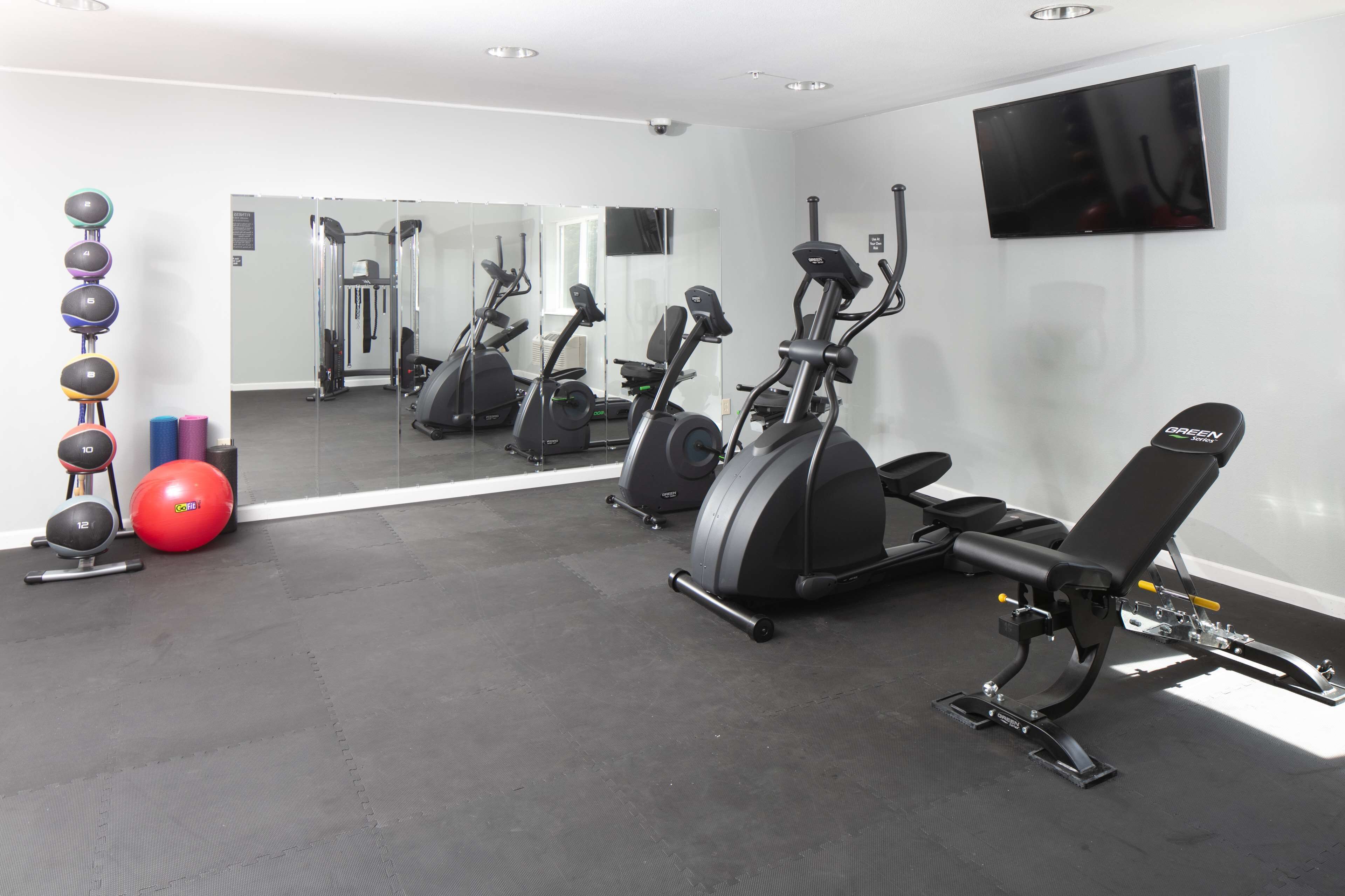 Une salle de sport équipée de matériel d’exercice, avec un téléviseur fixé au mur et un grand miroir qui reflète l’espace.