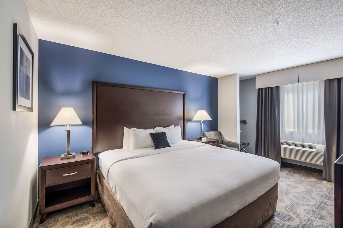 Une chambre d’hôtel avec un grand lit, deux tables de chevet avec des lampes, une chaise, un bureau et une fenêtre avec des rideaux.