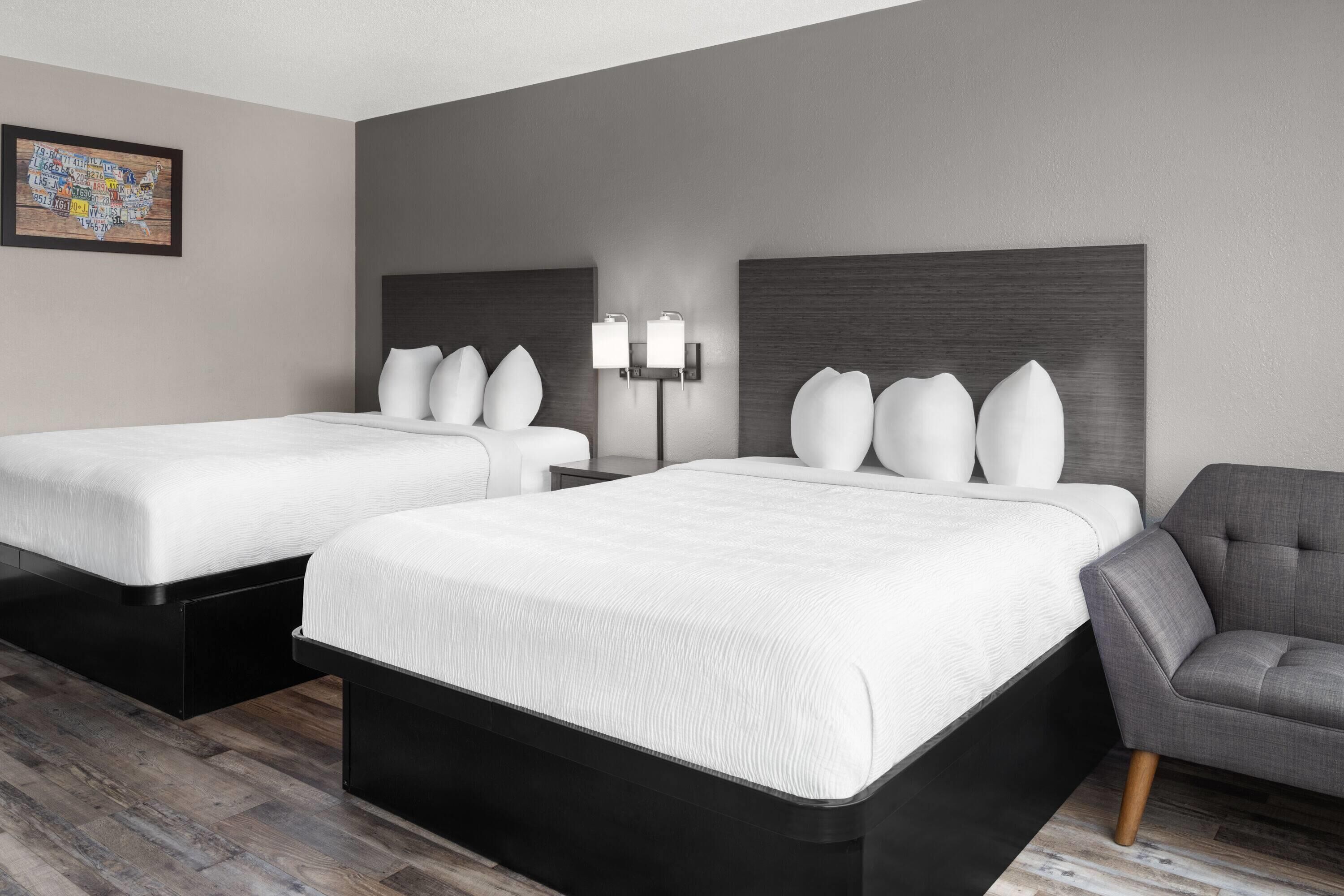 A hotel room with two beds, a gray armchair, and a wall-mounted lamp.