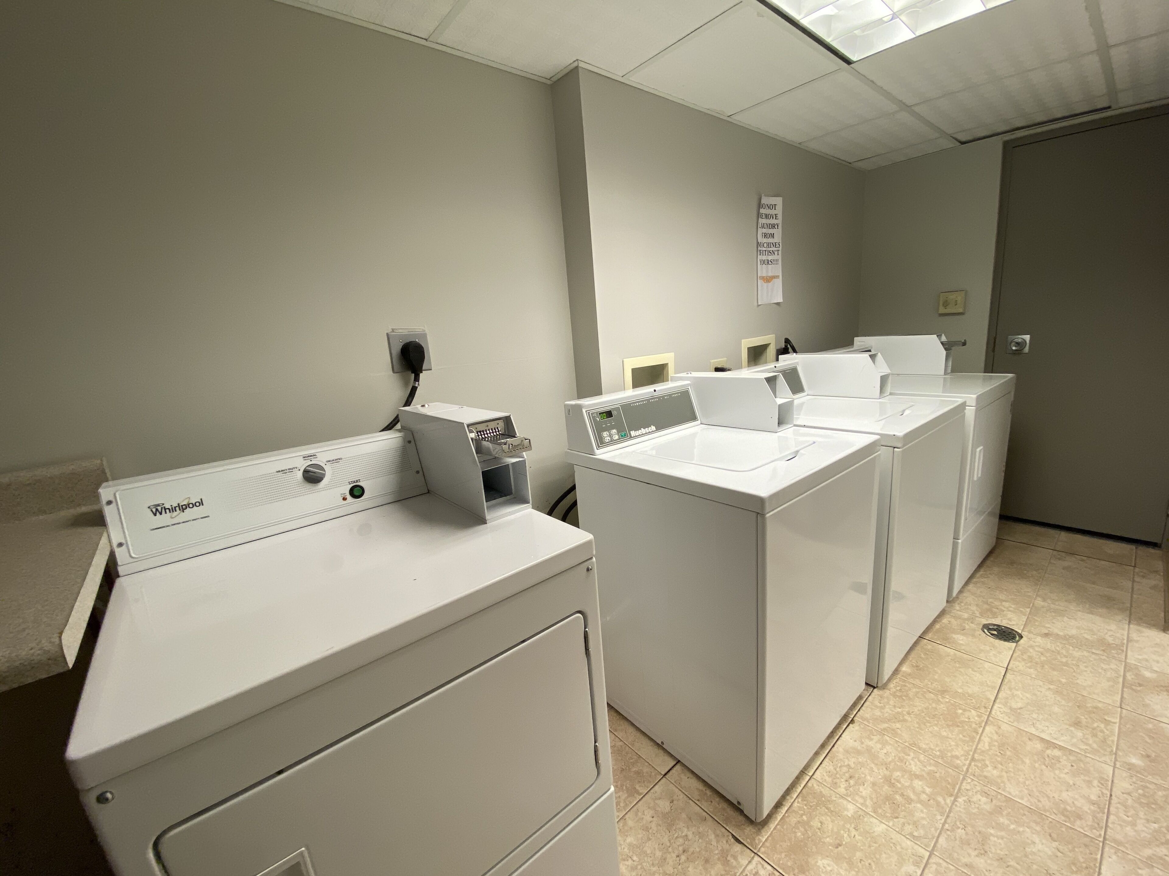 A laundry room with Whirlpool washers and dryers, a sign prohibiting removal of laundry from machines, and a tiled floor.