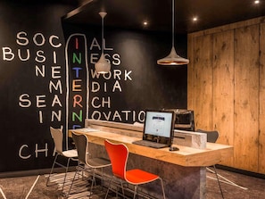 A modern workspace with a wooden desk, a laptop, and a computer monitor. There are two chairs, one red and one grey. The wall features a chalkboard with the words 'SOCIAL BUSINESS INTERNSHIP' written in white.