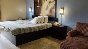 A hotel room with a bed, a chair, a bedside table with a phone, and a wall-mounted light.