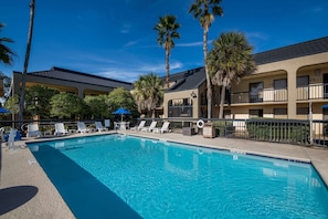 Une piscine entourée de palmiers et d’un bâtiment doté de balcons, avec des chaises longues et des parasols à proximité.