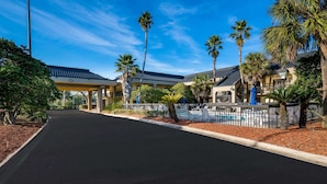A hotel with a covered walkway, palm trees, and a pool area.