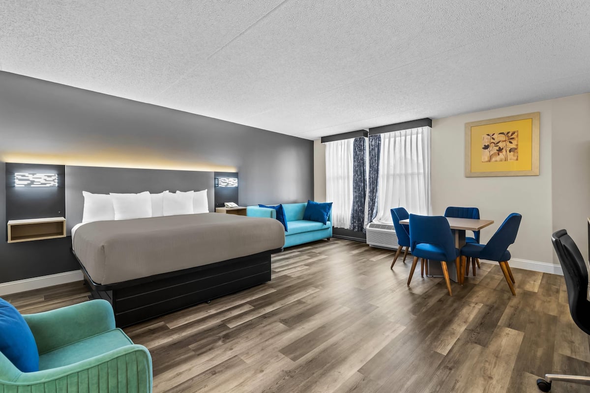 A modern hotel room with a large bed, a blue sofa, a dining table with chairs, and a small desk.