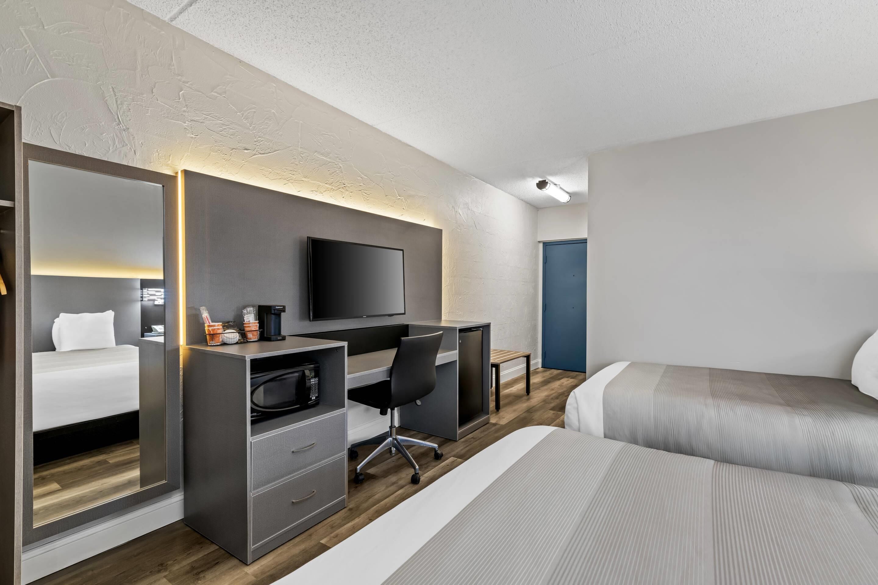 A modern hotel room with a bed, a desk with a computer, a television, and a mirror.