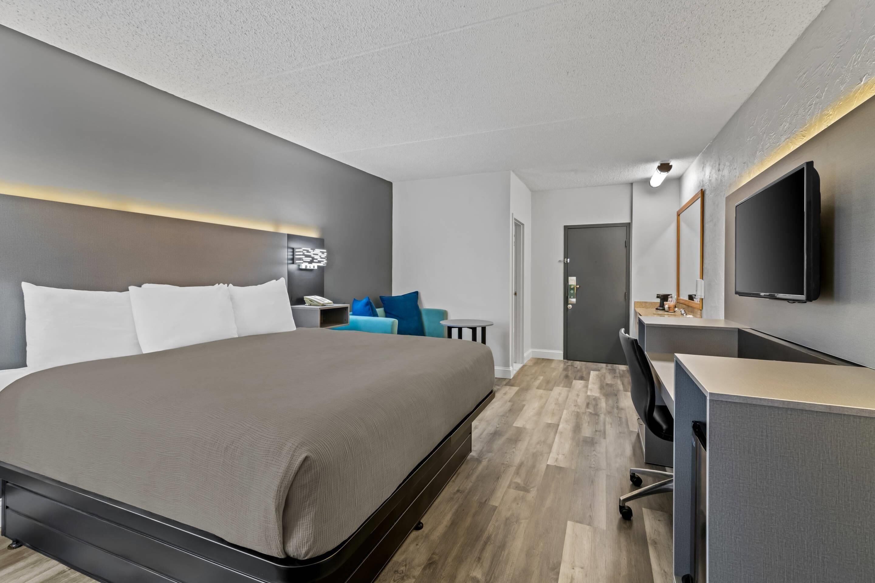 A modern hotel room with a large bed, a desk, a TV, and a seating area with a blue chair.