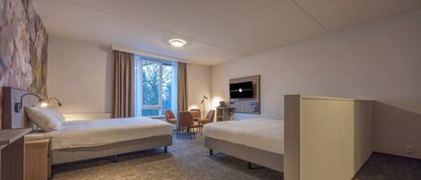 Ein Hotelzimmer mit zwei Einzelbetten, einem Schreibtisch mit Lampe, einem Stuhl, einem an der Wand befestigten Fernseher und einem großen Fenster mit Vorhängen.