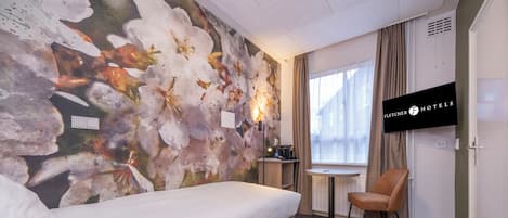 A hotel room with a bed, a desk with a chair, a TV, and a window with curtains.
