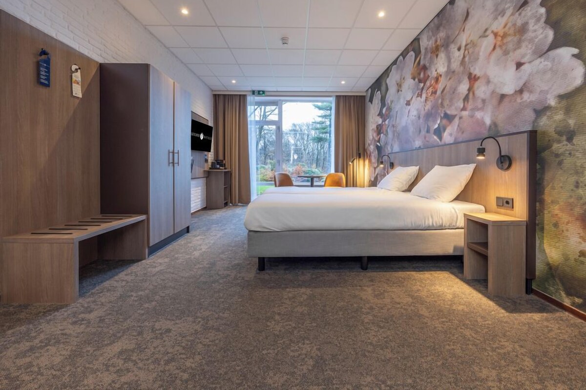 A hotel room with a bed, a desk, a chair, a bench, a wall-mounted TV, and a large window.