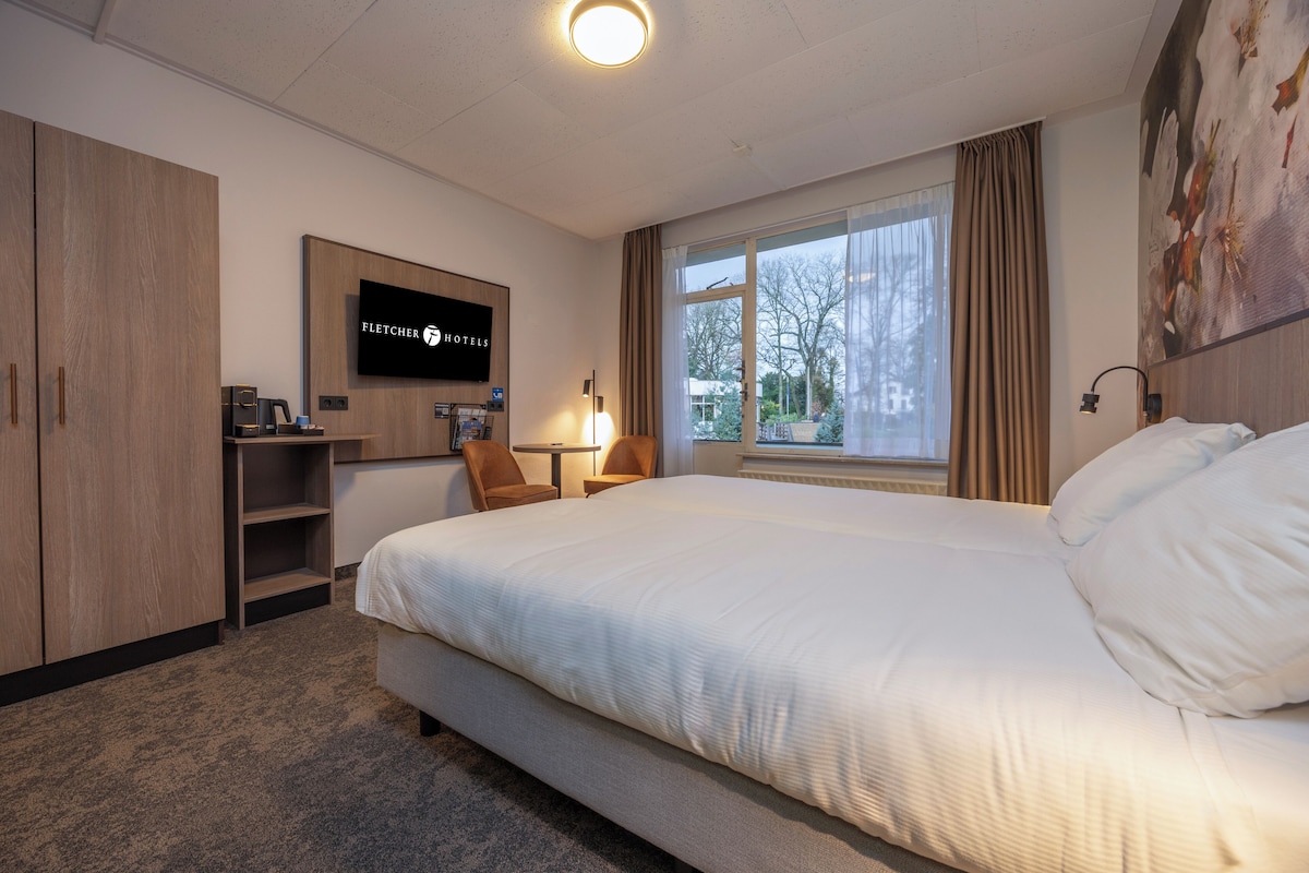 A hotel room with a large bed, a desk with a computer, a TV, and a window with curtains.