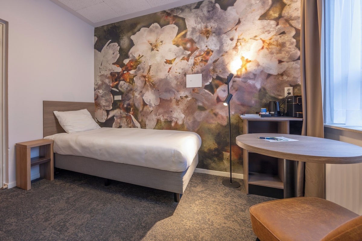 Ein Schlafzimmer mit einem Bett, einem Nachttisch, einem kleinen Schreibtisch mit Stuhl, einem runden Tisch und einer großen Blumenmalerei an der Wand.