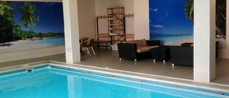 An indoor swimming pool with a view of a beach mural, a seating area with chairs and a sofa, and a wooden rack.