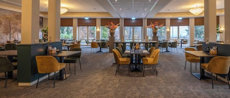 A spacious restaurant with high ceilings, large windows, and a carpeted floor. Tables and chairs are arranged for dining.