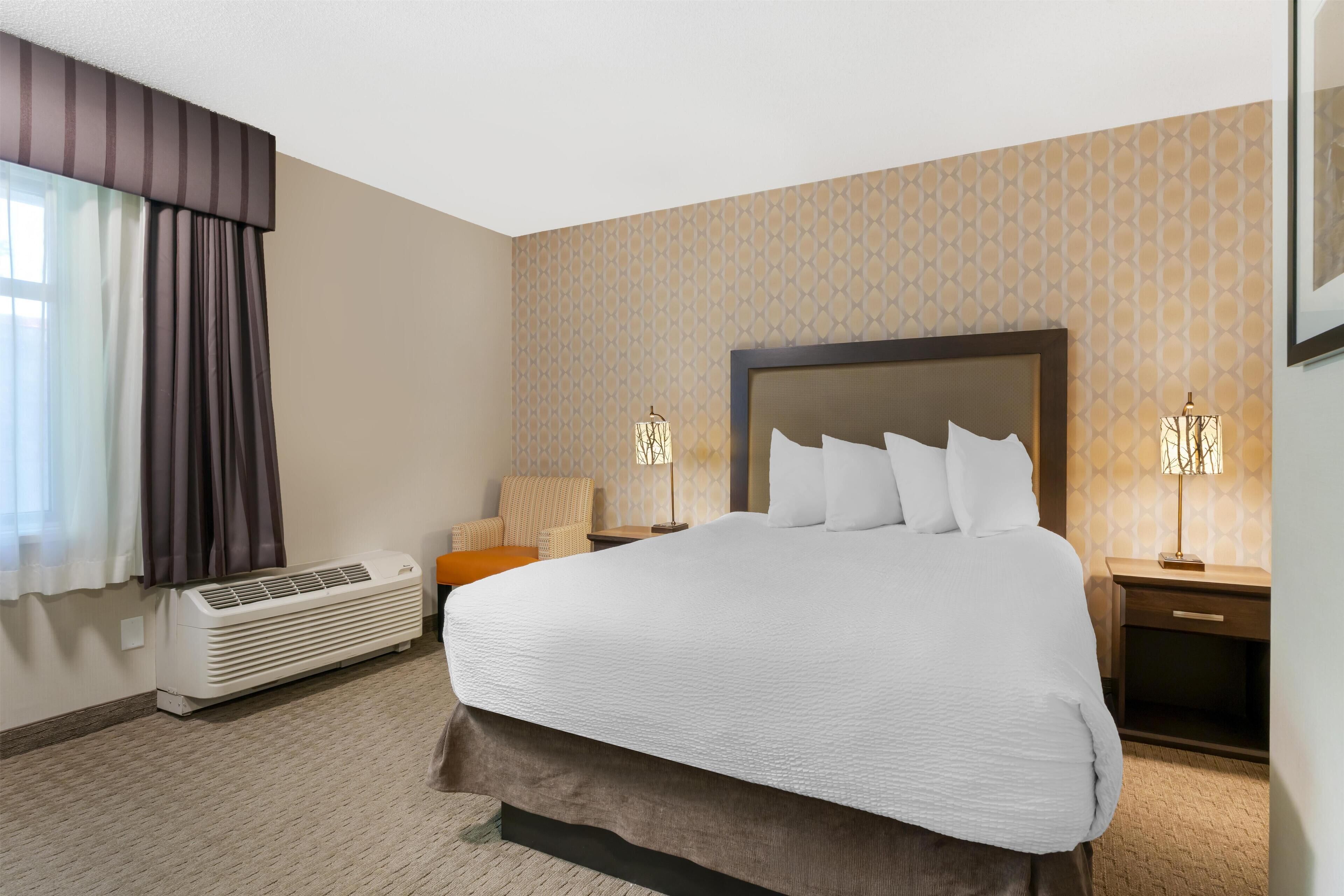 Quarto de hotel com uma cama grande, uma mesa de cabeceira com abajur, uma cadeira e ar condicionado fixado na parede.