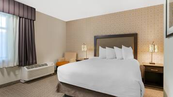 Quarto de hotel com uma cama grande, uma mesa de cabeceira com abajur, uma cadeira e ar condicionado fixado na parede.