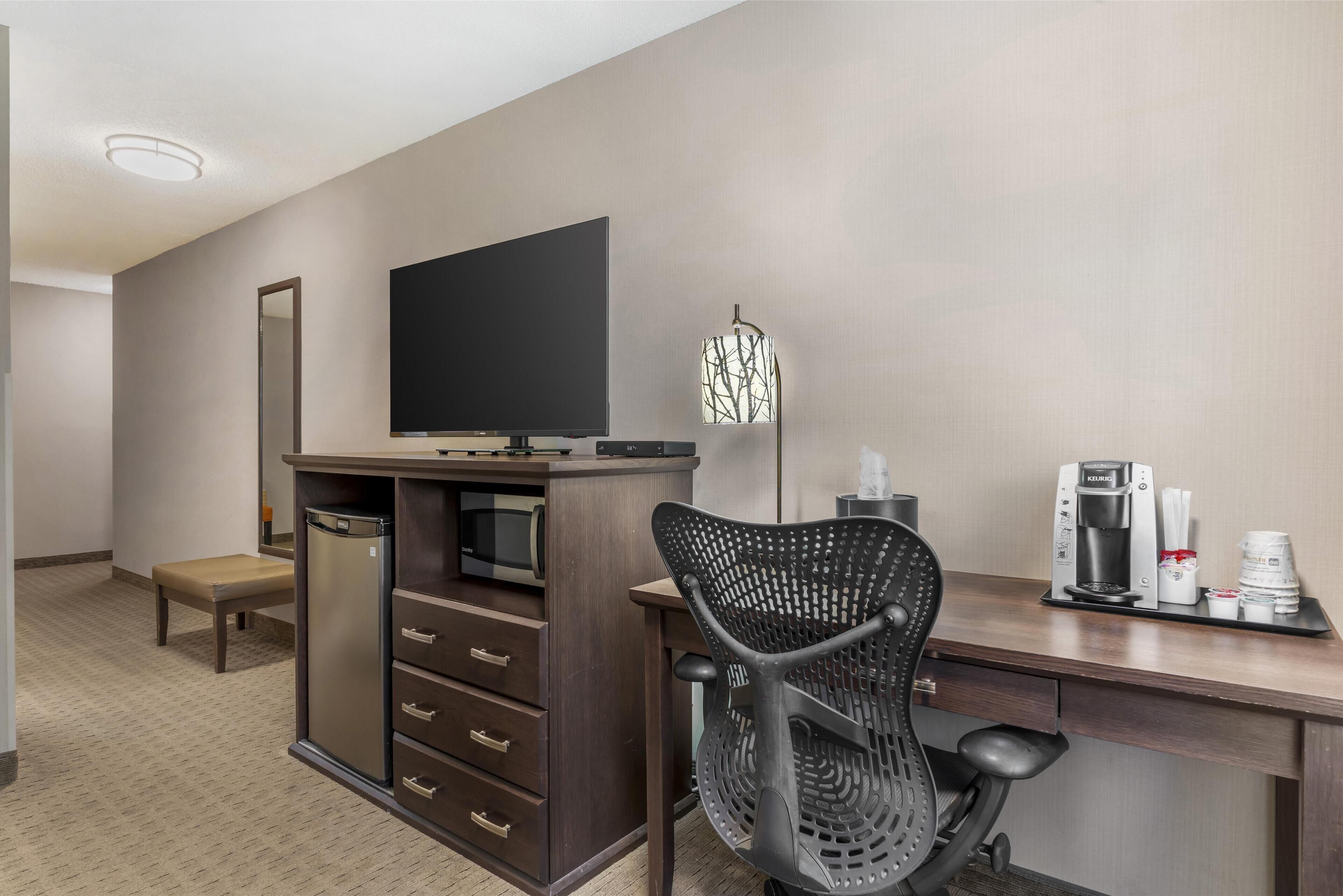A hotel room with a desk, chair, kitchenette, and a large flat-screen TV.
