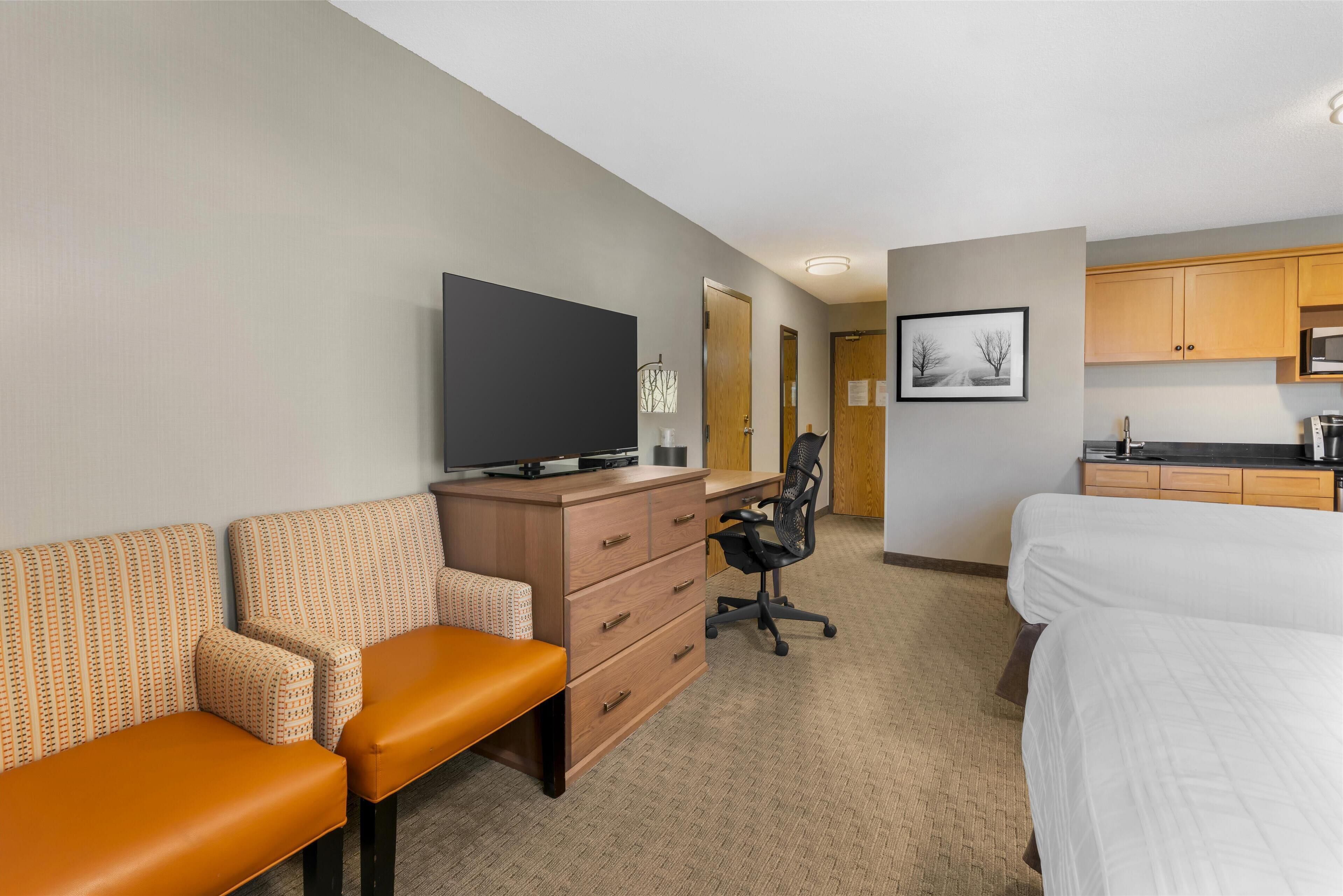 A hotel room with a bed, a desk with a chair, a television, a kitchenette, and a bathroom with a mirror.