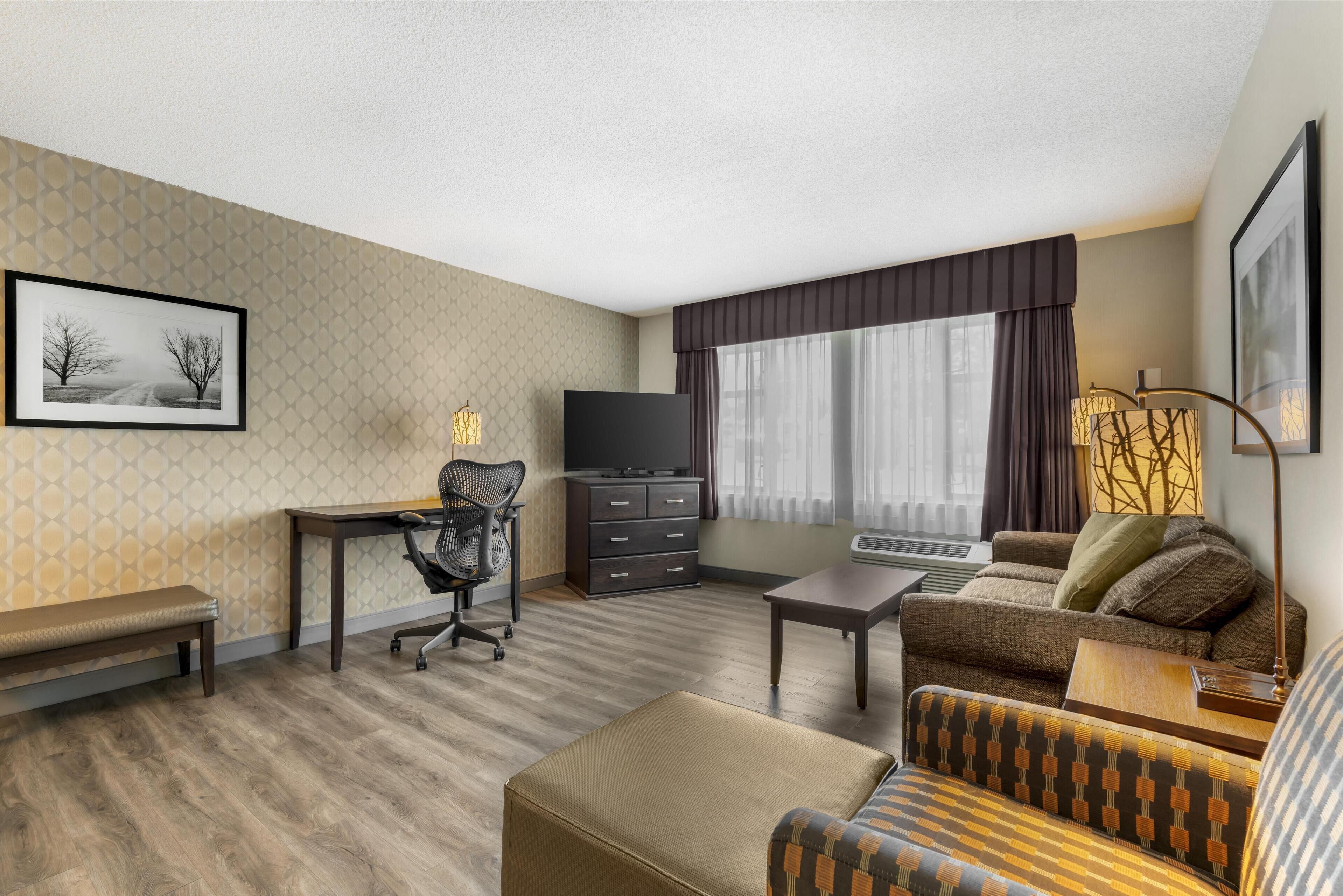 A hotel room with a sofa, a desk, a chair, a television, a bench, and a window with curtains.