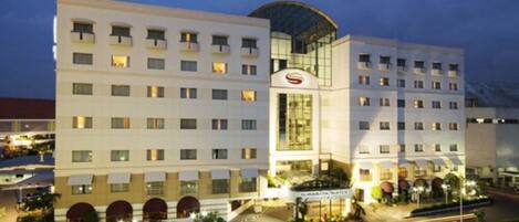 A multi-story hotel with a prominent glass facade, a central entrance with a sign, and a parking area in front.