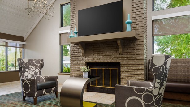 A modern living room with a fireplace, two patterned armchairs, a coffee table, and a flat-screen TV mounted on a brick wall. - Residence Inn by Marriott Orlando Altamonte Springs/Maitland (Altamonte Springs)