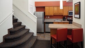 A compact kitchen with wooden cabinets, a stainless steel refrigerator, a microwave, and a sink. There are two red bar stools at a small island counter. - Residence Inn by Marriott Orlando Altamonte Springs/Maitland (Altamonte Springs)