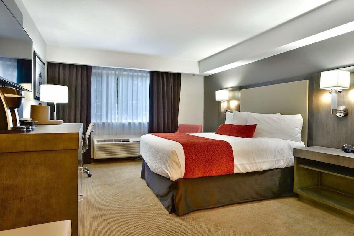 Quarto de hotel com cama, mesas de cabeceira, escrivaninha, cadeira, televisão e ar condicionado.