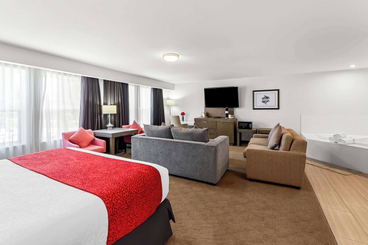 Quarto de hotel moderno com uma cama grande, um sofá cinzento, uma poltrona bege, uma secretária com candeeiro e uma televisão de ecrã plano.