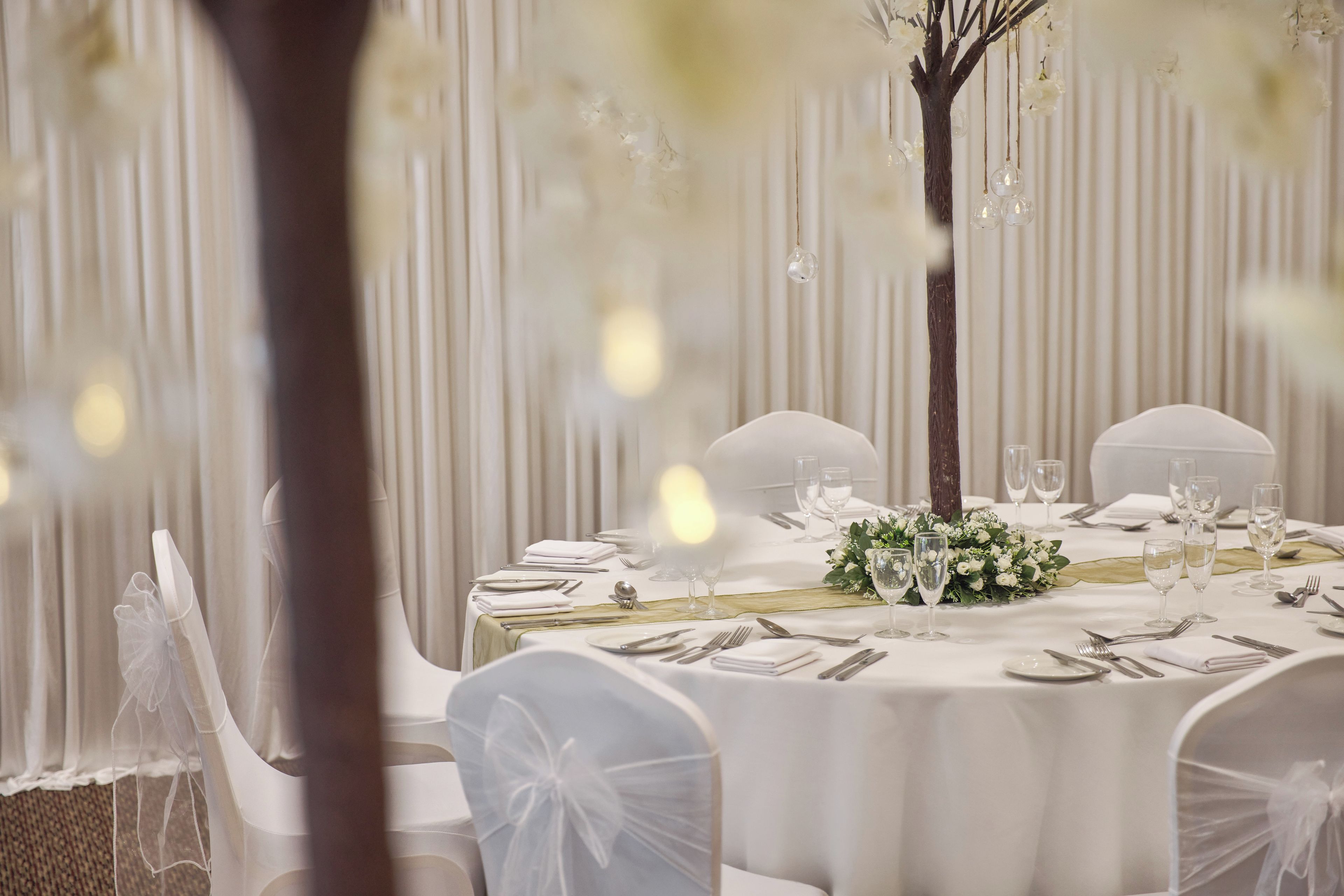 A formal dining table set with white tablecloth, glassware, and silverware, surrounded by chairs with white coverings.