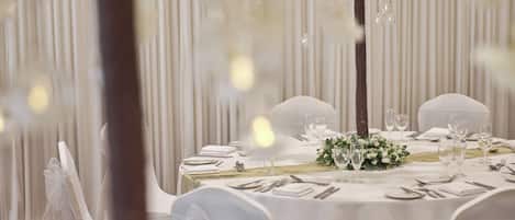 Mesa formal de comedor con mantel blanco, copas y cubiertos, rodeada de sillas con fundas blancas.
