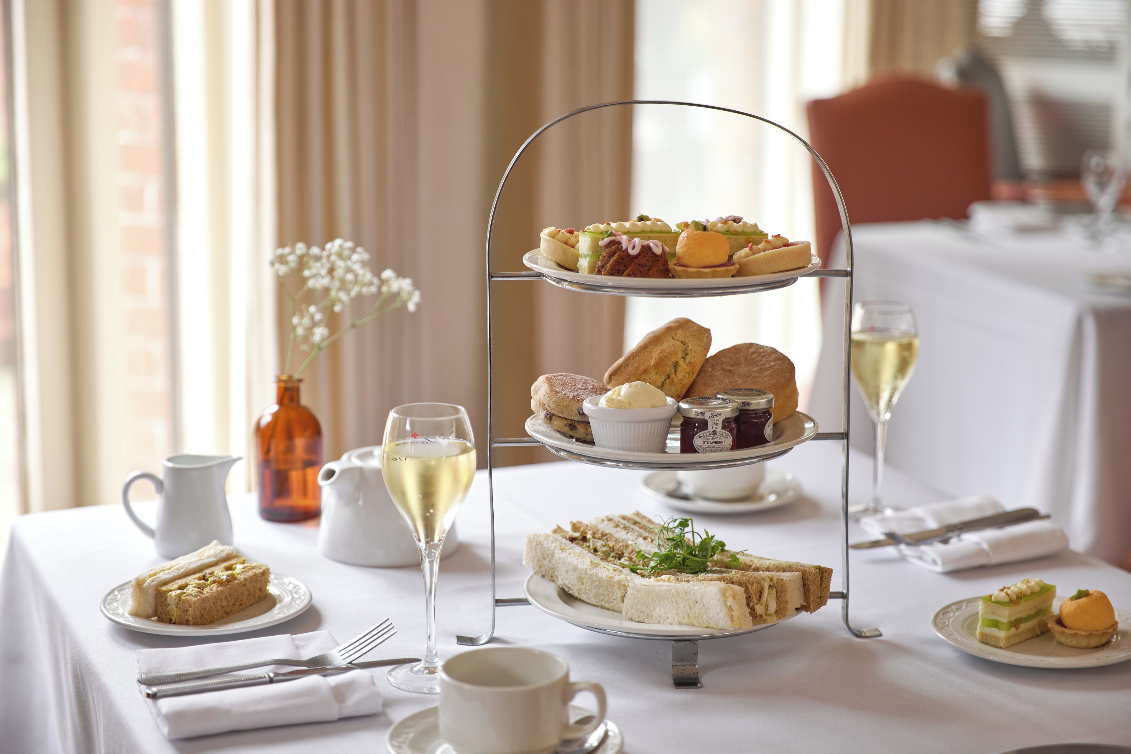 Ein dreistöckiges Kuchenregal mit verschiedenen Gebäcken und Sandwiches, ein Glas Weißwein und eine Vase mit Blumen auf einem Tisch.