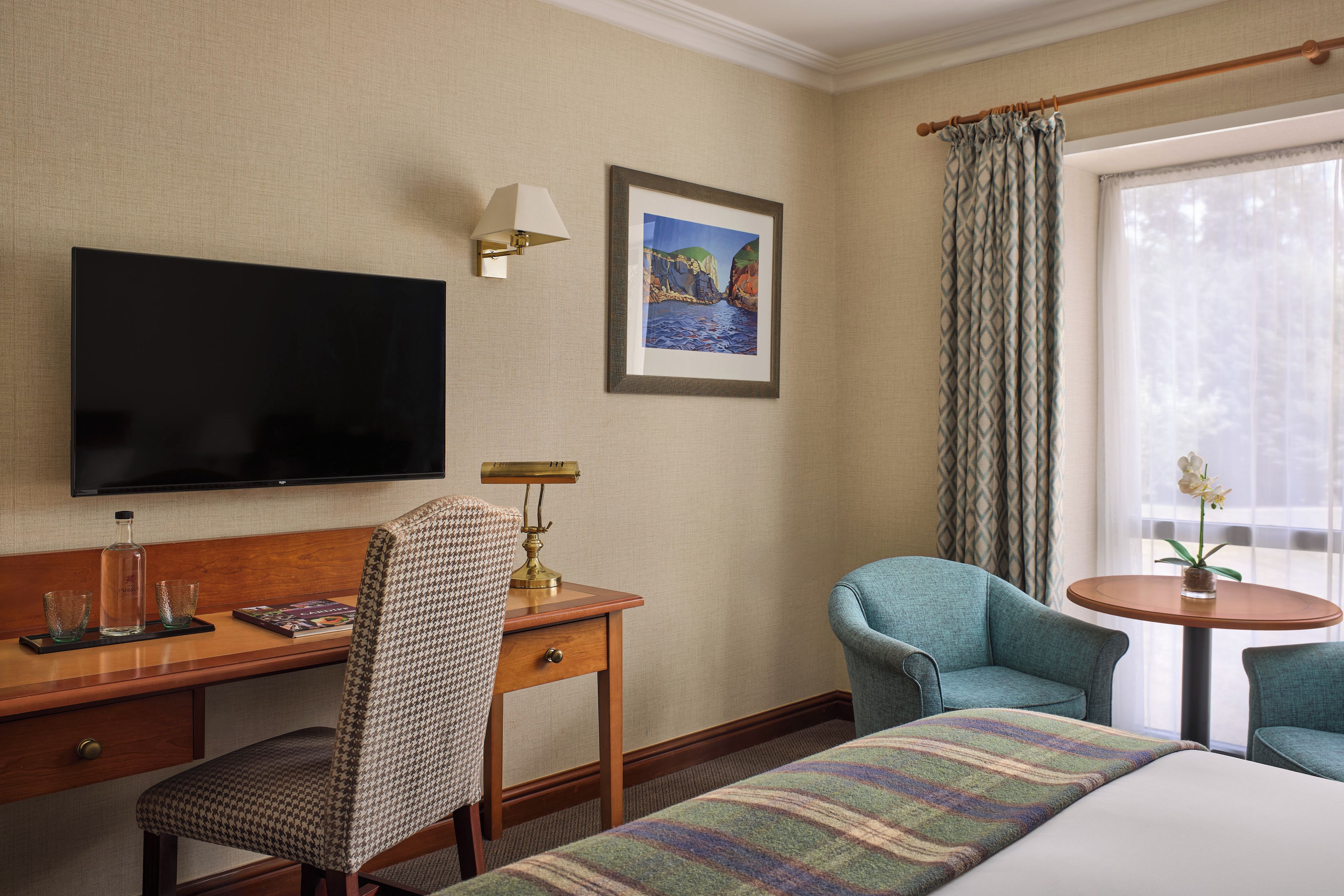 A hotel room with a bed, a desk with a chair, a television, a framed picture on the wall, and a window with curtains.