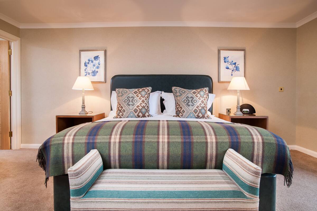 Una cama bien hecha con colcha a cuadros y almohadas a juego, flanqueada por dos mesitas de noche con lámparas y obras de arte enmarcadas en las paredes.