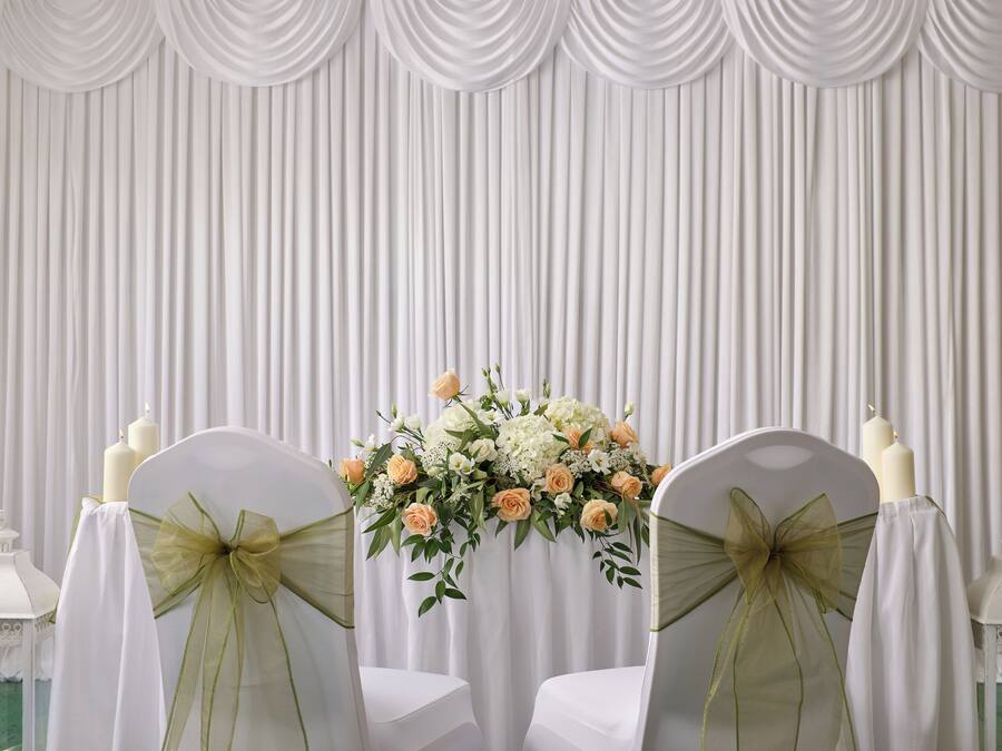 Una habitación con un fondo de cortina blanca, dos sillas con fundas blancas y lazos verdes, y un arreglo floral sobre una mesa.