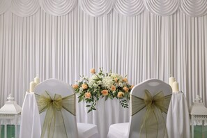 A room with a white curtain backdrop, two chairs with white covers and green bows, and a floral arrangement on a table. - Parkway Hotel & Spa (Cwmbran)