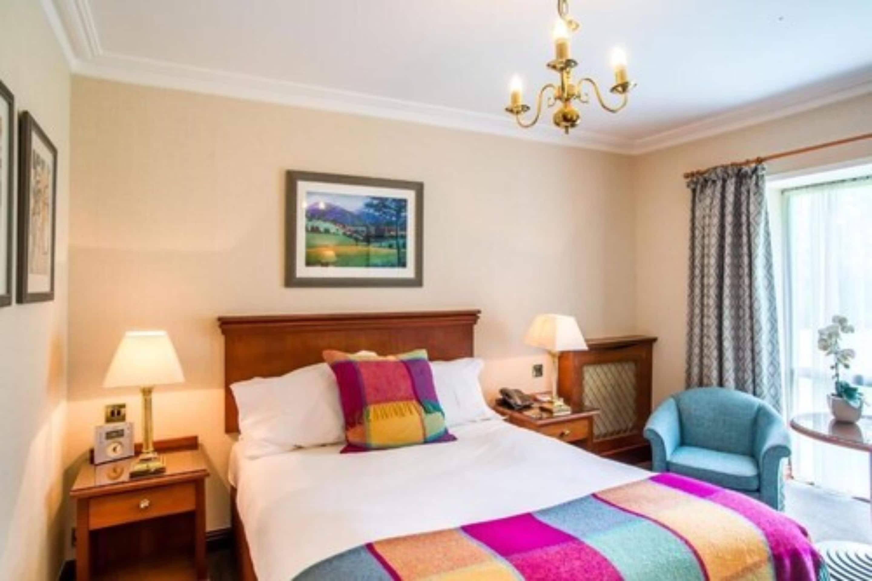 Una habitación de hotel con una cama, dos lámparas de noche, una mesita de noche, una silla, una ventana con cortinas y un candelabro.